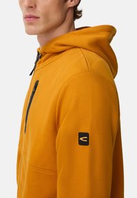 Homme portant un sweat à capuche jaune moutarde avec une poche zippée noire sur la poitrine et un petit patch logo noir sur la manche gauche.