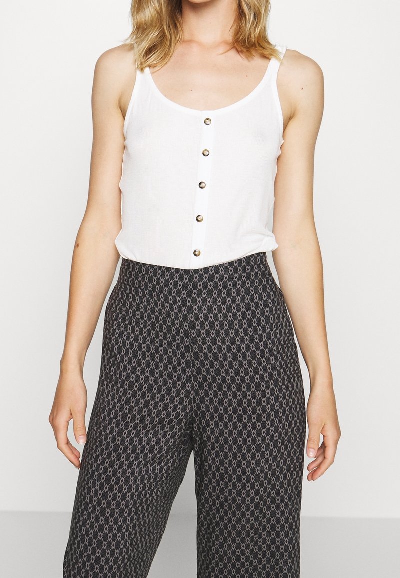 Femme portant un haut blanc sans manches avec des boutons et un pantalon noir taille haute avec un petit motif géométrique sur un fond uni.