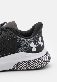 Zwarte sportieve schoen met een gestructureerde bovenkant, meshpatroon, wit logo en een witte tussenzool met "HOVR" branding. Rubberen buitenzool met gripontwerp.