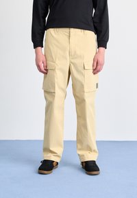 Beige cargo bukser med flere frontlommer og en afslappet pasform, lavet af letvægtsstof. Båret med sorte sko med brune detaljer.