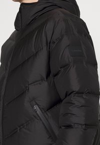 Giubbotto in piuma nero con tasca con zip, texture imbottita e cappuccio. Presenta un logo Calvin Klein sulla manica.