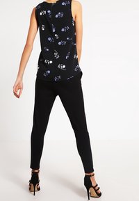 Donna che indossa un top floreale senza maniche nero, pantaloni neri aderenti e sandali con tacco alto neri, in piedi con la schiena rivolta verso l'osservatore.