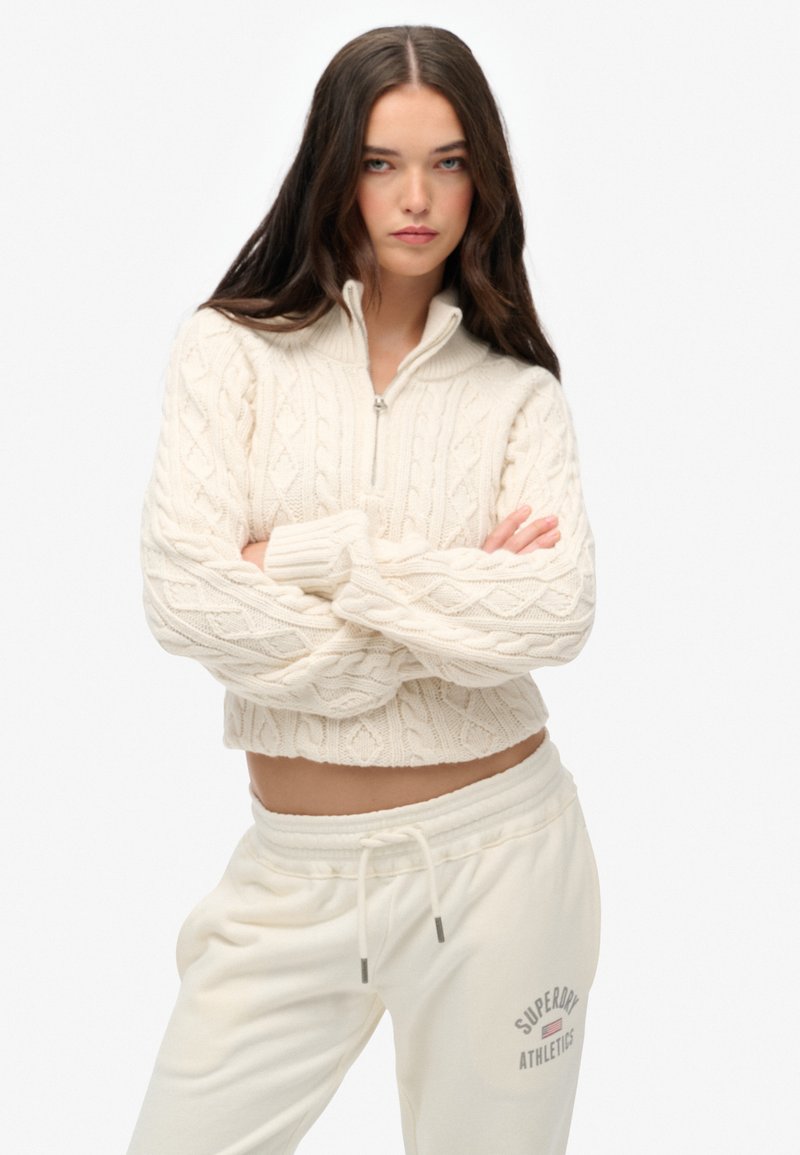 Crème kleur kabelgebreide cropped trui met een hoge kraag en halve rits, gecombineerd met bijpassende crème kleurige sweatpants met een logo op de heup.