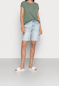 Grön kortärmad t-shirt, ljusblå denimshorts med fransade kanter, vita slidesandaler med logodetalj, stående på vit golv.