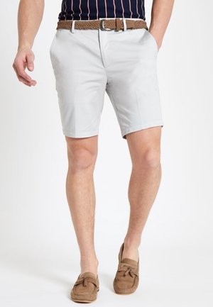 Homme portant un short gris clair, une ceinture tressée marron, une chemise à rayures bleu marine et des mocassins marron, debout les mains partiellement dans les poches.