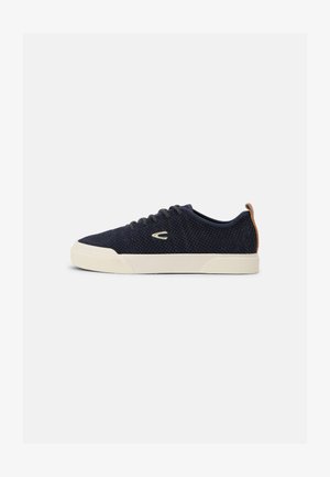 Navy mesh sneakers with round toe, white rubber sole, contrasting tan heel tab, and black laces; textured surface detail throughout.