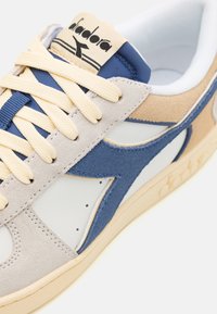 Adidași gri deschis și bej cu accente albastre. Prezintă materiale din suede și piele, șireturi plate bej și o talpă rotundă, texturată.