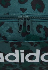 Grønn kamuflasjemønstret ryggsekk med doble grå glidelåsdragere og en stor hvit "adidas" logo på fronten. Teksturert stoff.