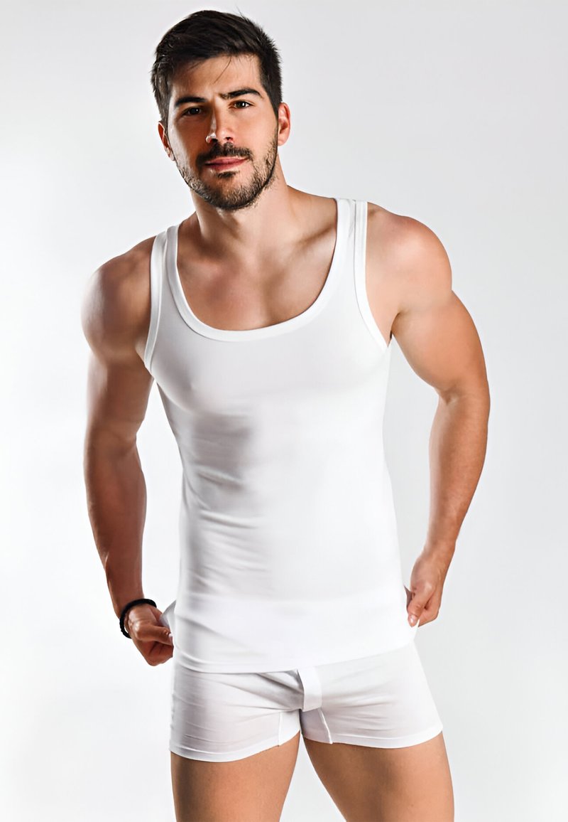 Man met kort donker haar en baard, gekleed in een wit mouwloos ondershirt en witte boxershorts, tegen een effen achtergrond.