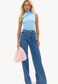 Femme blonde portant un haut bleu clair sans manches froncé, jean large bleu taille haute, tenant un sac à main rose duveteux, bracelet et bagues en or.