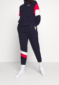 Tuta in pile blu navy con top a zip a quarto. Maniche a strisce bianche e rosse, pantaloni con vita elastica e sneakers bianche. Design casual e confortevole.