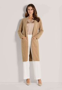 Frau in einem beigen langen, flauschigen Cardigan, taupefarbenem Top mit Spitzenbesatz, weißen weiten Hosen und beigen spitzen Pumps, die mit den Händen in den Taschen dasteht.