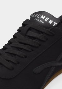 Baskets noires à lacets avec "PAVEMENT EST. 2010" imprimé sur la languette et un discret motif cousu sur le côté.