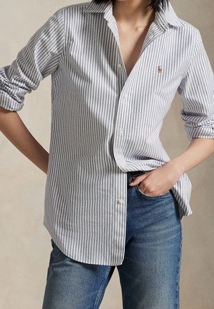 Persona che indossa una camicia a righe verticali azzurro chiaro e bianco con le maniche arrotolate e jeans blu, una mano in tasca.