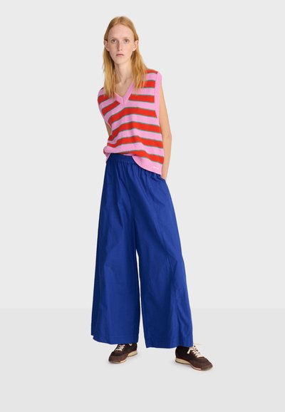 Junge Frau steht mit den Händen in den Taschen, trägt eine ärmellose Weste mit pinken und roten Streifen, weite blaue Hosen und braune Turnschuhe.