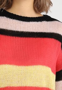 Dzianinowy sweter w poziome paski w kolorach czerwonym, żółtym, czarnym i różowym. Posiada luźny dekolt oraz lekką, teksturowaną tkaninę.