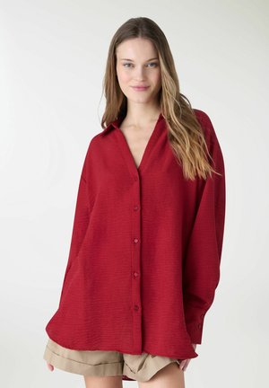 Giovane donna con capelli lunghi che indossa una camicia rossa oversize con bottoni e pantaloncini beige, in piedi davanti a uno sfondo bianco semplice.