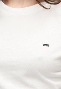 Κοντινό σε ένα άτομο που φοράει απλό λευκό μπλουζάκι με στρογγυλή λαιμόκοψη και μικρό ορθογώνιο κόκκινο, λευκό και μπλε λογότυπο στο στήθος.