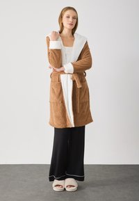 Brown, fluffy robe with a white sherpa-lined hood, two patch pockets, a tie belt, worn over a white top and black pants, paired with fluffy slippers.