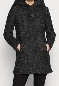 Cappotto nero texturizzato con cappuccio, dotato di una linea dritta, due tasche laterali e un motivo a pois sottile su tutto il tessuto.