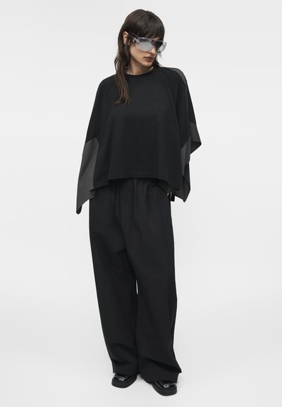 Γυναίκα με μαύρο υπερμεγέθες τοπ με φαρδιά μανίκια, φαρδιά μαύρη παντελόνα, μαύρα παπούτσια και μεγάλα διαφανή γυαλιά ηλίου, στέκεται μπροστά σε λευκό φόντο.