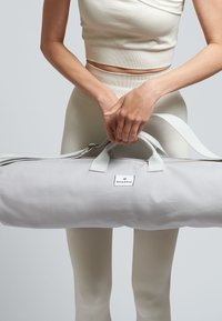 Bolsa de yoga de color gris claro hecha de lona, con dos asas, cierre de cremallera y un parche de logo visible. Textura suave, forma cilíndrica.
