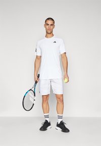 Weißes Sportshirt mit schwarzen Akzenten, weiße Shorts, schwarze Turnschuhe und gestreifte Socken. Hält einen Tennisschläger und einen gelben Tennisball.