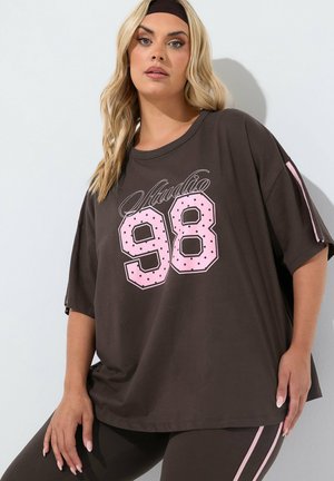 Mujer con cabello rubio largo que lleva una cinta negra en la cabeza y una camisa negra de gran tamaño con estampado rosa "Studio 98" y leggings negros a juego con rayas rosas.
