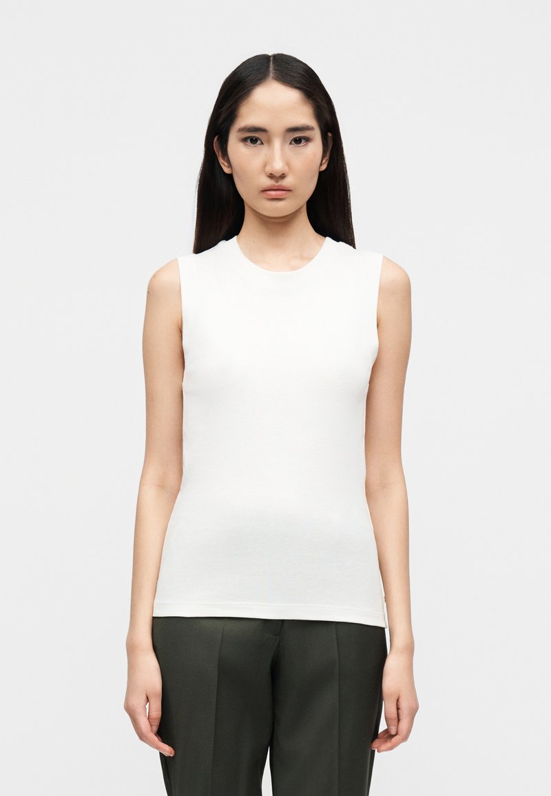 Λευκή αμάνικη μπλούζα από βαμβάκι, με στρογγυλή λαιμόκοψη και εφαρμοστή σιλουέτα, συνδυασμένη με σκούρο πράσινο παντελόνι.