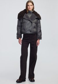 Kobieta w czarnej skróconej kurtce puchowej z owczym kołnierzem, szarym swetrze z golfem, czarnych szerokich spodniach i brązowych masywnych butach.