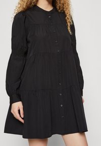 Black button-up dress featuring a tiered design, long puff sleeves, and a collared neckline. Made from smooth, lightweight fabric.