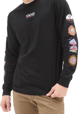 Černé triko s dlouhým rukávem značky Vans s barevnými psychedelickými symboly na rukávech, oblečené osobou v béžových kalhotách, ruka spočívá u kapsy.