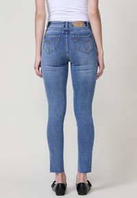 Jean en denim bleu ajusté avec un ourlet effiloché, taille haute, et poches arrière avec des coutures décoratives. Texture lisse.