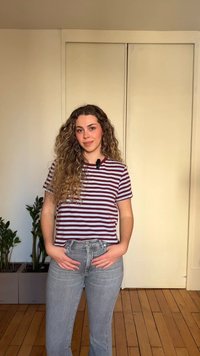 Γυναίκα με μακριά σγουρά μαλλιά, ντυμένη με ριγέ μπλουζάκι και γκρι τζιν, στέκεται μέσα στον χώρο με τα χέρια στις τσέπες, φυτά και κλειστές πόρτες πίσω της.