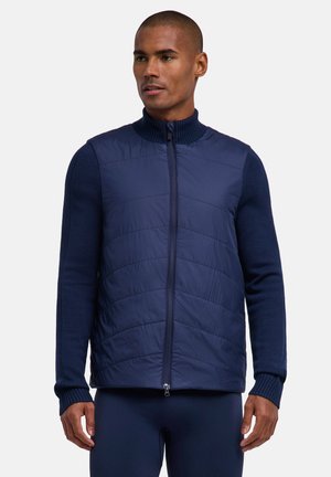 Mann trägt eine marineblaue gesteppte Zip-up-Weste über einem marineblauen Strickoberteil mit langen Ärmeln, steht mit entspannten Händen seitlich vor grauem Hintergrund.