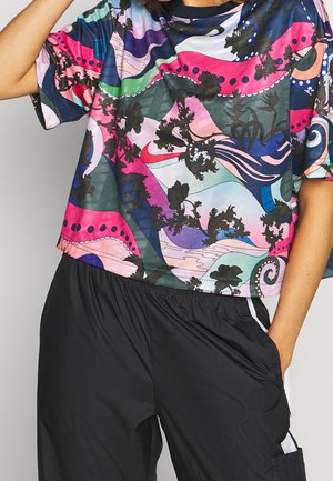 Osoba ubrana w koszulę z abstrakcyjnym wzorem w wielokolorowe motywy i luźne czarne spodnie, z rękami częściowo w kieszeniach, na jednolitym tle.