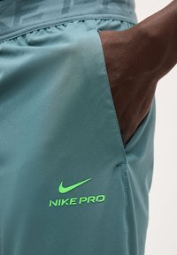 Χέρι στην τσέπη βερμούδας Nike Pro σε τιρκουάζ χρώμα με φωτεινό πράσινο λογότυπο στο μηρό, που δείχνει την ελαστική ζώνη μέσης και την απαλή υφή του υφάσματος.