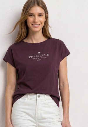 Uśmiechnięta kobieta w ciemnofioletowej koszulce z napisem „Palm Club” i białych spodniach stoi na tle jednolitej jasnej ściany.