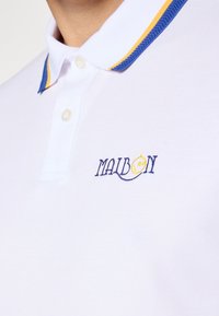 Hvit polo skjorte med en blå og gulstripet krage, med en marineblå brodert logo "MAIBON." Stoffet ser glatt og pustende ut.
