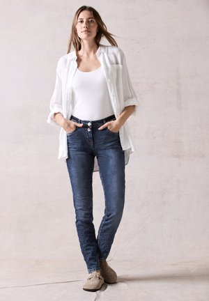 Skinny jeans van donkerblauw denim met een licht vervaagd effect, gecombineerd met een witte tanktop en een losse, doorschijnende witte blouse.