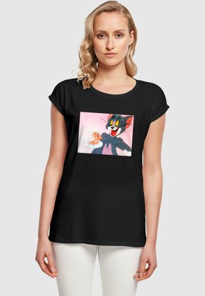 Svart T-skjorte med et fargerikt grafisk motiv av en tegneserie-katt og mus, med løs passform og korte ermer. Designet har en rosa bakgrunn.
