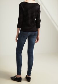 Svart tröja med trekvartsärmar och ett strukturerat mönster, ihop med åtsittande blå denimjeans och svarta slip-on skor med guldaccenter.