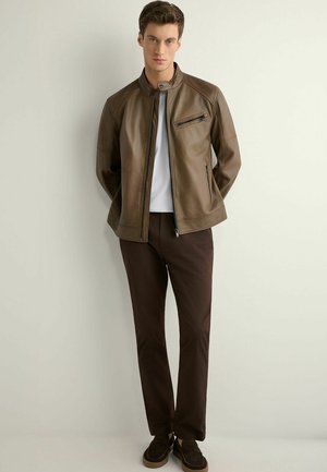 Młody mężczyzna stoi oparty o ścianę, ubrany w brązową skórzaną kurtkę, białą koszulę, ciemnobrązowe spodnie i ciemne mokasyny, z rękami za plecami.