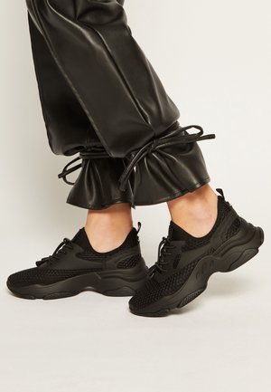 Piedi che indossano sneakers chunky nere e pantaloni in pelle neri con polsini con coulisse, visibili su uno sfondo semplice.