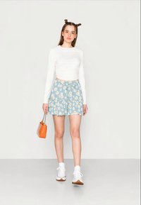 Blusa leve de mangas compridas e uma saia azul com padrão floral e detalhe em laço, combinada com ténis brancos e uma mala laranja.