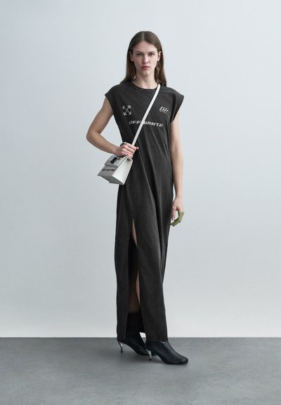 Γυναίκα που φοράει μαύρο μακρύ φόρεμα χωρίς μανίκια με πλαϊνή σχισμή, κρατάει μια λευκή τσάντα χιαστί και φοράει μαύρα μποτάκια αστραγάλου σε γκρι φόντο.