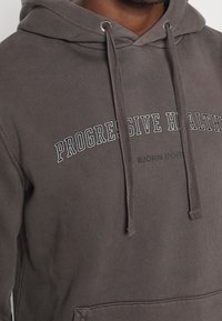 Gros plan sur une personne portant un sweat à capuche gris foncé avec le texte "PROGRESSIVE HEALTH" et des cordons visibles à l'avant.