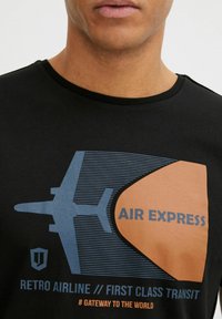 Camiseta de algodón negra con un gráfico de un avión azul y un fondo naranja, que incluye el texto "AIR EXPRESS" y "RETRO AIRLINE // FIRST CLASS TRANSIT."