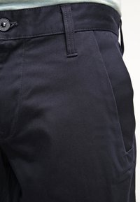 Närbild av mörkblå byxor i midjan med synlig bältesöglor, knapp och sidoficka på en ljus bakgrund.