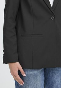 Črn blazer s enojnim gumbom, zareznimi klapnicami in stranskim žepom. Teksturirana tkanina in prilagojen kroj, v kombinaciji z modrimi jeans hlačami.